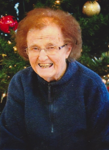 Carol smiles, looking like she is laughing. She wears a blue sweater in front of a Christmas tree.