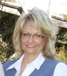 Sheryll smiles. She is standing outside and wearing a white button-up and blue vest.