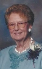 Iona smiles. She wears a blue embroidered sweater in front of a blue backdrop with white earrings, necklace, and rose pinned to her lapel.